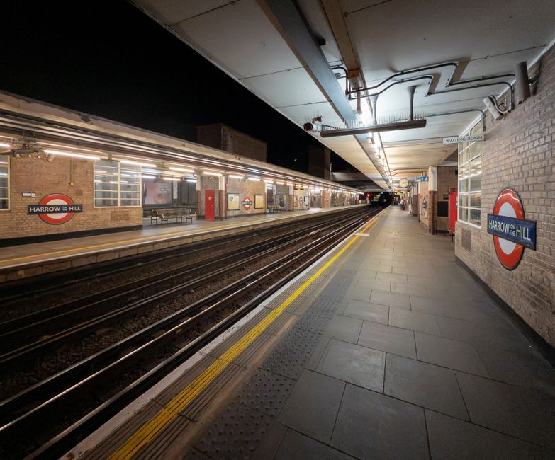 Interior view of Harringay station platform during evening or nighttime, featuring brick walls and various signage including a digital display indicating train information, with a staircase leading to